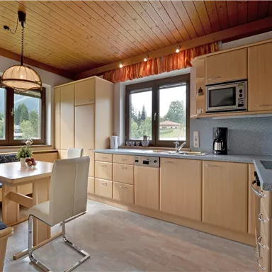 Eine moderne Küche mit Holzdecke und großen Fenstern. Der Essbereich hat einen Tisch und Stühle, sowie eine Küchenzeile mit verschiedenen Geräten.