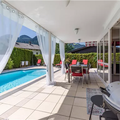 A modern terrace overlooking a pool and beautiful mountain views. There is a dining area with red chairs and a grill.