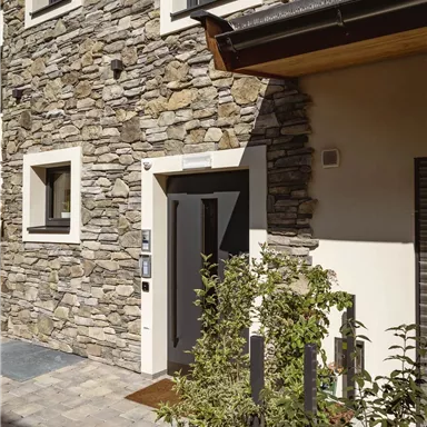 A modern house with a stone facade and large windows. The entrance area is adorned with plants.