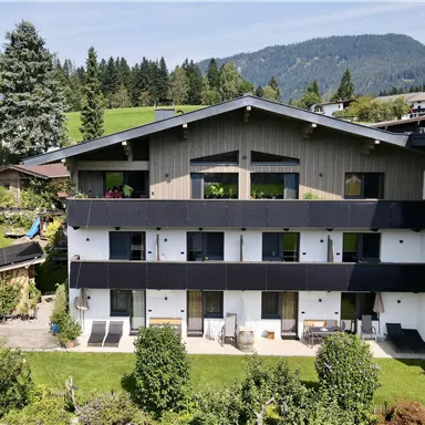 Ein modernes Gebäude umgeben von grünen Wiesen und Bäumen. Im Hintergrund sind Berge und ein blauer Himmel zu sehen.