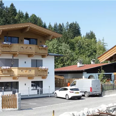 A modern building with wooden cladding and balconies stands next to a traditional wooden house. The surroundings are surrounded by trees and the sky is clear.