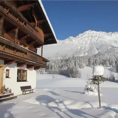 Ein gemütliches Chalet im Winter, umgeben von viel Schnee. Im Hintergrund erheben sich majestätische Berge unter einem klaren Himmel.