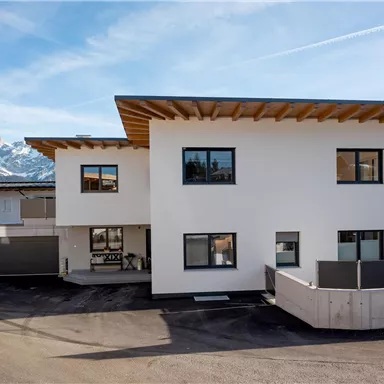 Ein modernes Gebäude mit weißer Fassade und Holzdetails. Im Hintergrund sind schneebedeckte Berge zu sehen.
