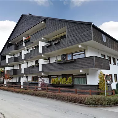 Ein modernes Wohngebäude mit dunklem Holzdesign und weißen Wänden. Die Umgebung ist grün mit Sträuchern und Bäumen.