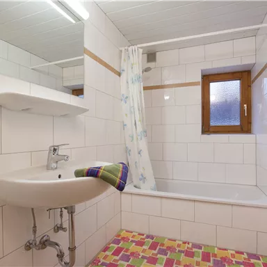 Ein einfaches Badezimmer mit einer Wanne, Waschbecken und einem Fenster. Die Wände sind gefliest und der Boden hat ein buntes Muster.