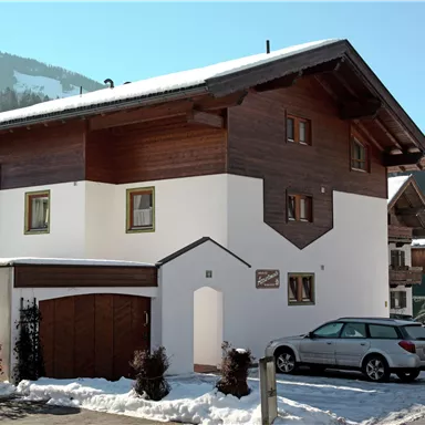 Ein modernes Haus mit Holzverkleidung, umgeben von Schnee. Im Vordergrund steht ein Parkplatz mit einem Auto.