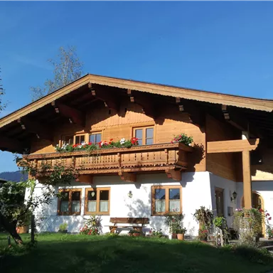 A traditional alpine house with wooden cladding and a large balcony. Surrounded by green areas and a clear blue sky.