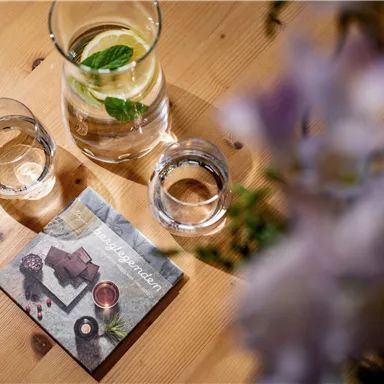 Ein Tisch mit einem Krug Wasser, zwei Gläsern und einer Menükarte. Im Hintergrund sind Blüten zu sehen.