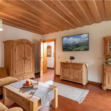 Ein gemütlicher Wohnraum mit Holzverkleidung und einer hellen Einrichtung. Eine Couch, ein Tisch mit Obst und ein TV-Bildschirm im Hintergrund sind sichtbar.