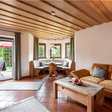 Gemütliches Wohnzimmer mit Holzbalkendecke und einem großen Fenster. Es gibt eine bequeme Couch und einen Esstisch mit Bänken.