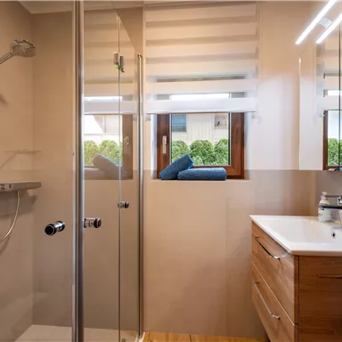 Ein modernes Badezimmer mit einer Glasdusche und einem Holzwaschtisch. Das Fenster bietet einen Blick auf einen begrünten Außenbereich.