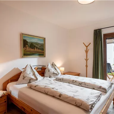 Ein gemütliches Schlafzimmer mit zwei Betten, bunten Kissen und einem Gemälde an der Wand. Große Fenster mit Blick auf den Garten sorgen für Tageslicht.