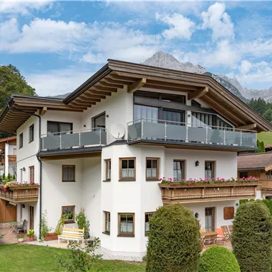 Ein modernes Haus in ländlicher Umgebung, umgeben von Bäumen und Bergen. Der Himmel ist leicht bewölkt, was eine ruhige Atmosphäre schafft.