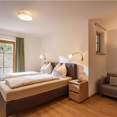 Ein modernes Schlafzimmer mit einem Doppelbett und zwei Kissen. Es gibt einen gemütlichen Sessel und große Fenster mit Vorhängen.