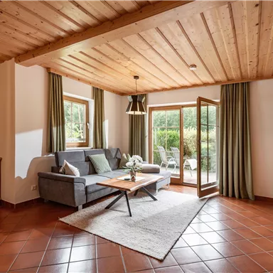 Ein gemütliches Wohnzimmer mit einem grauen Sofa und einem Holztisch. Große Fenster bieten Blick auf den Garten.
