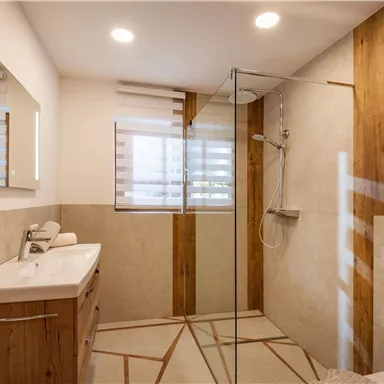 Ein modernes Badezimmer mit Glasdusche und Holzdetails. Die Wände sind licht und freundlich gestaltet, mit einem großen Spiegel über dem Waschbecken.