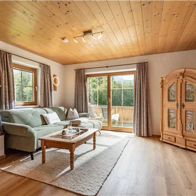 Ein gemütliches Wohnzimmer mit einem Holzofen, einer Sofalandschaft und einem großen Fenster. Helle Holzdetails und ein Teppich schaffen eine einladende Atmosphäre.