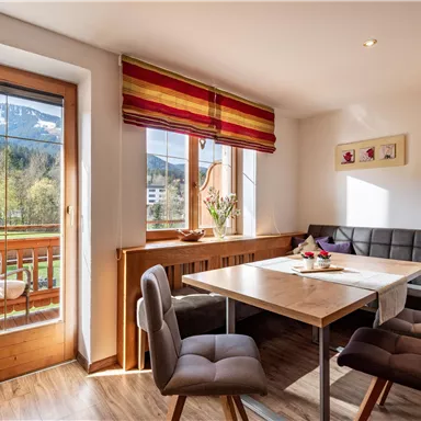 A cozy dining table in a bright room with a view of the mountains. Large windows let in plenty of light and showcase a balcony outside.
