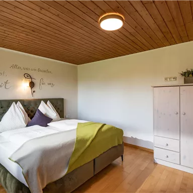 A cozy bedroom with a large bed and a modern wooden wall. Next to the bed stands a white wardrobe and a decorative wall clock.
