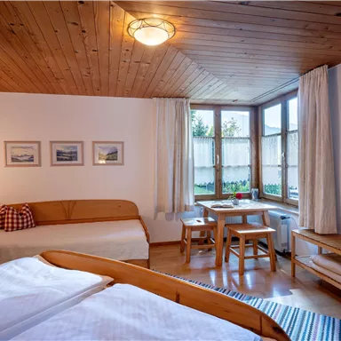Ein gemütliches Zimmer mit Holzdecke und großen Fenstern. Es gibt zwei Betten, eine Sitzecke und einen kleinen Tisch.