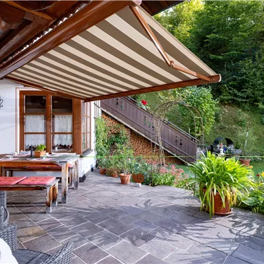 Eine gemütliche Veranda mit Gartenmöbeln und einem Essplatz. Im Hintergrund ist ein schöner Garten mit einer Treppe und vielen Pflanzen zu sehen.