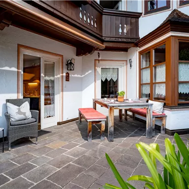 Eine gemütliche Terrasse mit einem Holztisch und Stühlen. Die Umgebung ist grün und freundlich gestaltet.