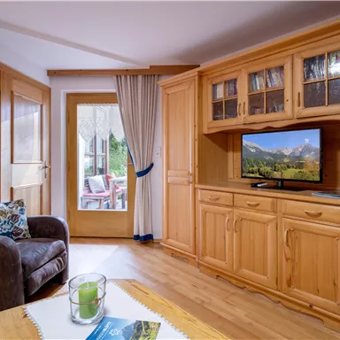 Ein gemütliches Wohnzimmer mit einem bequemen Sessel und Holzregalen. Ein Fernseher zeigt eine Landschaft, während ein Fenster den Blick nach draußen ermöglicht.