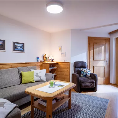 Ein gemütliches Wohnzimmer mit einem grauen Sofa und einem Holzkaffee Tisch. An den Wänden hängen Bilder, und ein Fenster lässt Tageslicht herein.