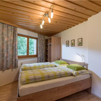 Ein gemütliches Schlafzimmer mit einem Doppelbett und bunten Kissen. Große Fenster sorgen für viel natürliches Licht und einen Blick ins Grüne.