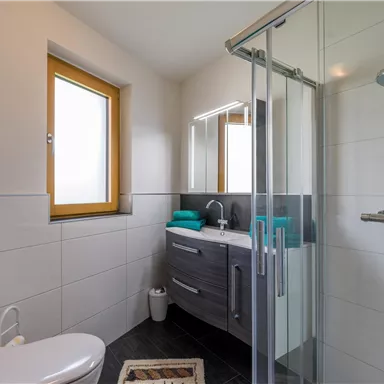 Ein modernes Badezimmer mit Dusche, Waschbecken und wandhängendem WC. Die Farben sind hell und freundlich, mit Handtüchern in Türkis.