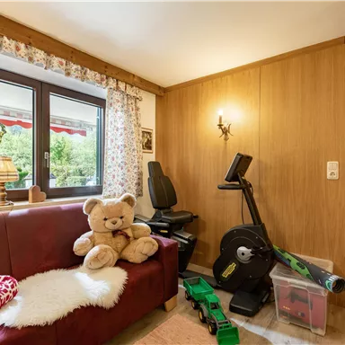 Ein gemütlicher Raum mit einem roten Sofa und einem großen Stofftier. Im Hintergrund befinden sich ein Fitnessgerät und dekorative Vorhänge.