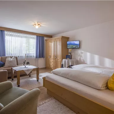 Ein gemütliches Zimmer mit einem großen Bett und einer sitzenden Couch. Helle Farben und viel Tageslicht schaffen eine einladende Atmosphäre.