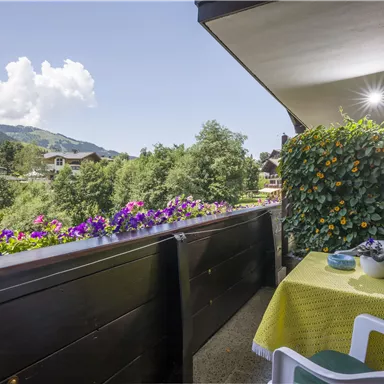 Ein gemütlicher Balkon mit einem schönen Ausblick auf Berge und Bäume. Ein Tisch mit gelber Tischdecke und einer Blumenkiste sorgt für eine einladende Atmosphäre.