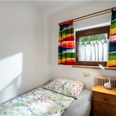 A small, bright room with a single bed and colorful curtains. On the nightstand, there is a lamp and the window lets in plenty of light.