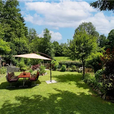 A beautiful garden with green grass and many trees. In the foreground, there is a table and chairs under a sunshade.