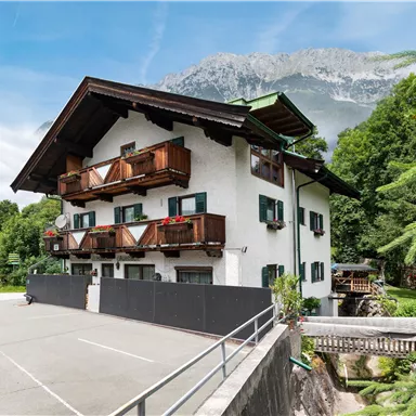 A pretty multi-story house with a balcony and a green environment. In the background, mountains and a blue sky can be seen.