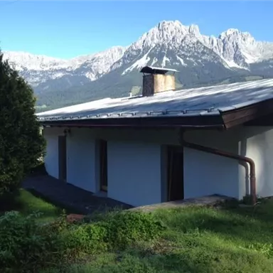 A modern house in a green environment with a view of majestic mountains. The sky is clear and sunny.