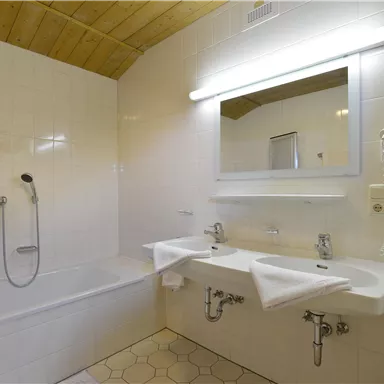 A modern bathroom with a bathtub and two sinks. It has bright tiles and a large mirror light.