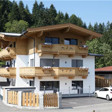 A modern building in alpine style with wooden balconies. Surrounded by green trees and a parking lot.