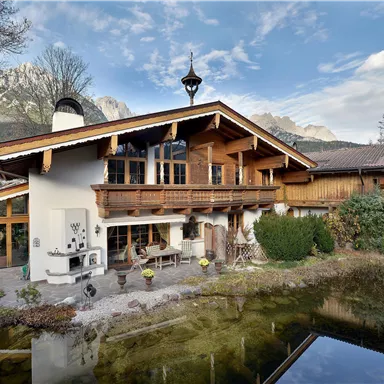 Ein schönes Holzhaus im Alpenstil mit großen Fenstern und einem gepflegten Garten. Im Vordergrund ist ein kleiner Teich zu sehen.