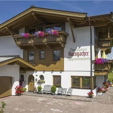 Ein alpenländisches Gebäude mit mehreren Balkonen, geschmückt mit bunten Blumen. Im Vordergrund befinden sich Parkplatz und Sitzgelegenheiten.