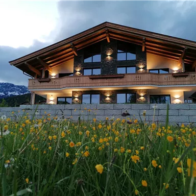 Ein modernes Ferienhaus in den Bergen, umgeben von bunten Blumen. Der Himmel ist bewölkt und die alpine Landschaft ist sichtbar.