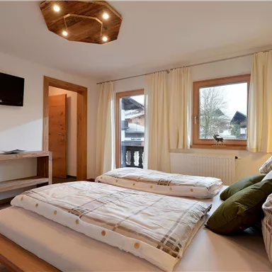 Ein gemütliches Schlafzimmer mit zwei Einzelbetten und großen Fenstern. An der Wand hängt ein Fernseher, und der Raum hat eine helle, freundliche Atmosphäre.
