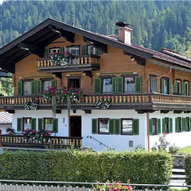Ein traditionelles, mehrstöckiges Holzhaus mit grünen Fensterläden und Blumenschmuck. Umgeben von einer gepflegten Hecke und der malerischen Alpenlandschaft.