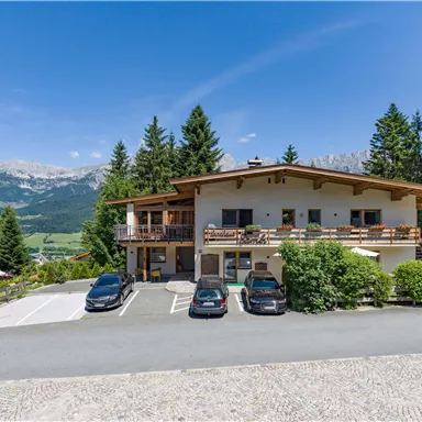 A building in alpine style, surrounded by trees and mountains. In front of the house are several cars parked on a paved lot.