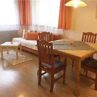 A bright living room with a cozy sofa and a table with chairs. Large windows allow plenty of light in, and the orange decoration creates an inviting atmosphere.
