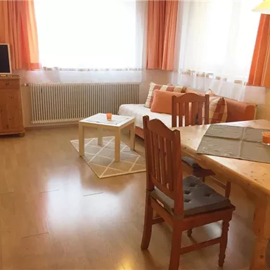 A cozy living room with a television, a table, and chairs. Large windows with orange curtains provide plenty of natural light.