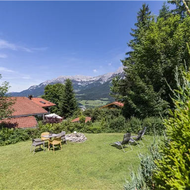 An idyllic landscape with a green garden and a view of the mountains. In the foreground, there are deck chairs and a fire pit.