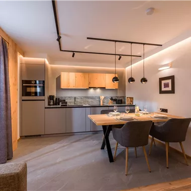 A modern kitchen with a dining area. The room is bright and inviting, furnished with gray chairs and wooden details.