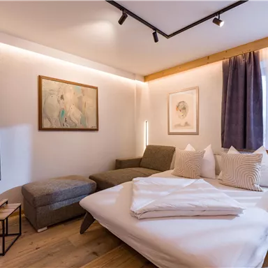 A modern room with a comfortable bed and a cozy seating area. Pictures hang on the wall and the room has large windows with curtains.